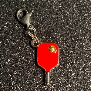 以下が含まれることがあります： 黄色いボールが付いたミニチュアのピクルボールラケットがついた赤と銀のキーホルダー。キーホルダーには銀色のロブスタークラスプが付いています。