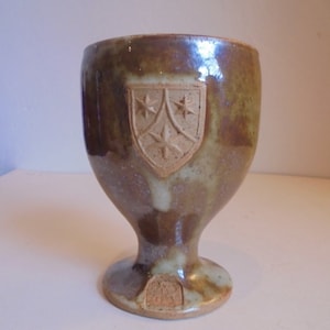 May include: A brown ceramic goblet with a white glaze and a raised shield design featuring three stars. The goblet has a wide base and a rounded body.