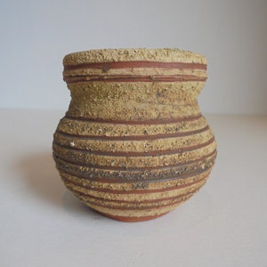 May include: A brown and tan ceramic vase with a textured surface and horizontal lines. The vase has a rounded body and a wide opening.