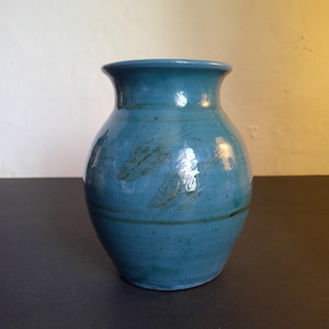 May include: A teal blue ceramic vase with a rounded body and a slightly flared opening. The vase has a subtle glaze and a few brown streaks.