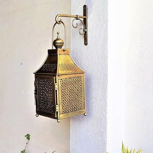 May include: A gold metal lantern with intricate designs hangs from a wall bracket. The lantern has a square shape with a pointed top and a decorative handle. The lantern is surrounded by green plants.
