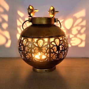 May include: A gold metal candle lantern with a decorative cut-out design and two bird finials. The lantern is lit with a candle inside.