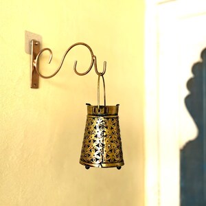 May include: A gold metal wall-mounted lantern with a decorative floral design. The lantern is hanging from a curved metal bracket.