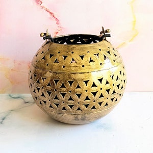 May include: A gold metal lantern with a decorative cut-out design. The lantern has two handles on the top and is round in shape.