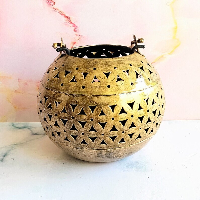 May include: A gold metal lantern with a decorative cut-out design. The lantern has two handles on the top and is round in shape.