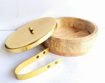 Puede incluir: Un cuenco de madera redondo con una tapa dorada y un asa de metal dorado. El cuenco está hecho de madera clara y tiene un acabado natural. La tapa tiene un pequeño pomo en la parte superior.