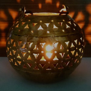 May include: A gold metal candle lantern with a decorative cut-out design of flowers and geometric shapes. The lantern is round and has a handle on each side.