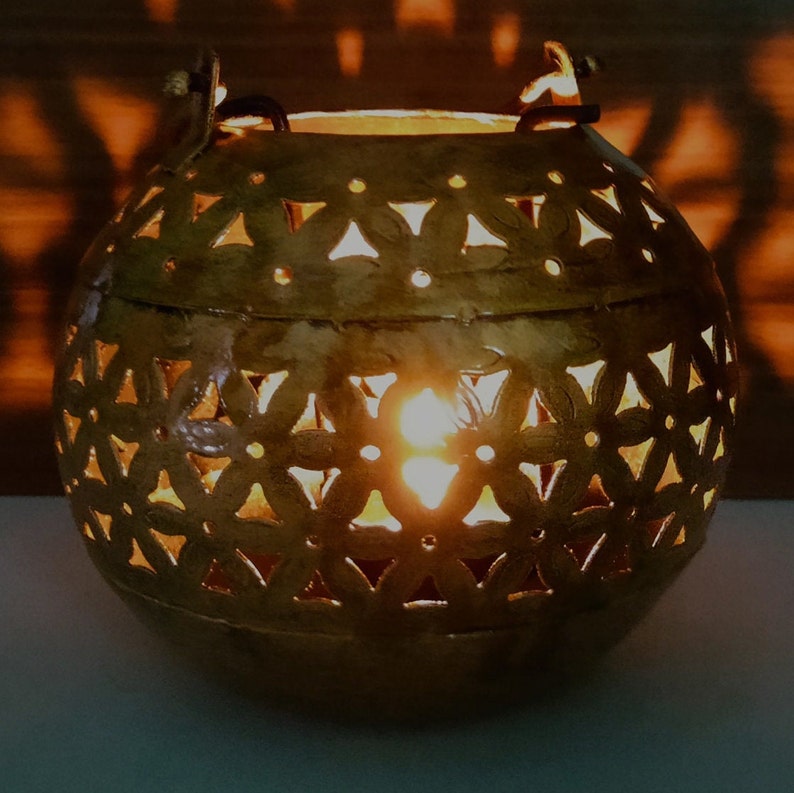 May include: A gold metal candle lantern with a decorative cut-out design of flowers and geometric shapes. The lantern is round and has a handle on each side.