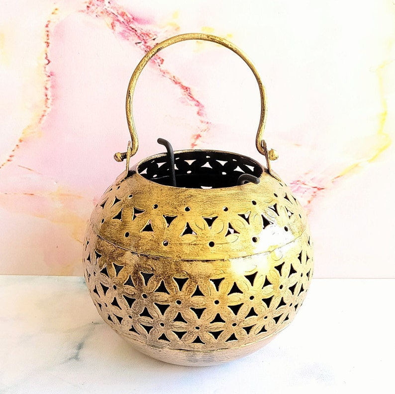 May include: A gold metal incense burner with a decorative floral pattern and a handle. The burner is round and has a wide opening at the top.