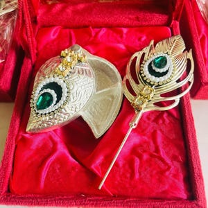 May include: A silver and gold jewelry set with a peacock feather design, featuring a large green gemstone and white pearl accents. The set is displayed in a red velvet box.