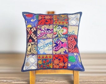 May include: A square patchwork pillow with a blue background and colorful floral patterns. The pillow is made of different fabrics and has a variety of textures. It is sitting on a wooden stand.