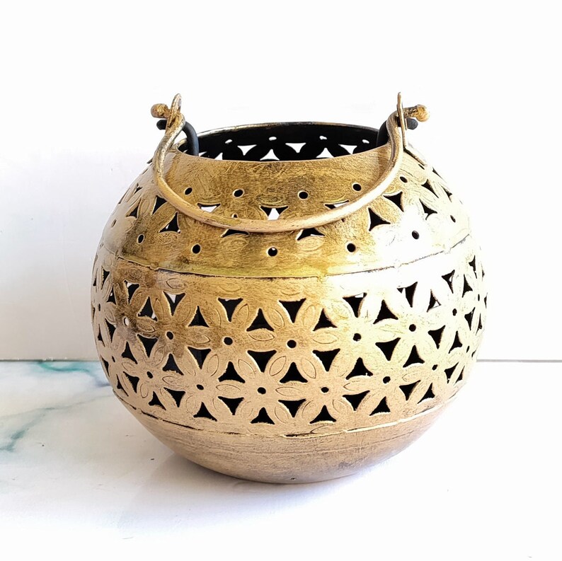 May include: A gold metal candle lantern with a round shape and a cut-out design of flowers. The lantern has a black metal handle.
