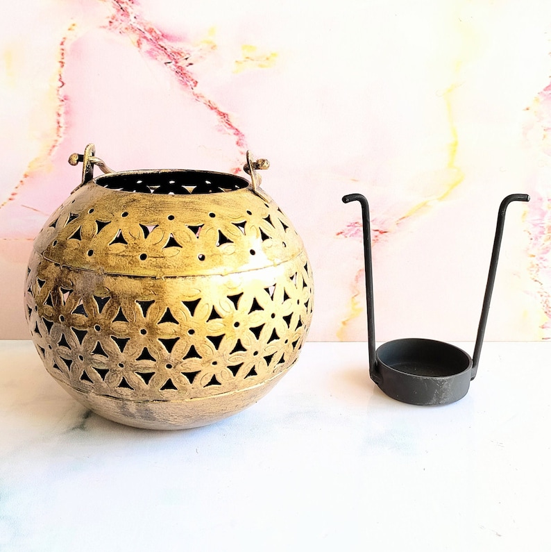 May include: A gold metal candle lantern with a decorative floral pattern. The lantern has a handle and is sitting on a white surface. A black metal candle holder is also shown in the image.