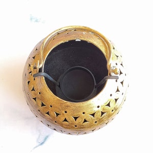 May include: Gold metal candle holder with a decorative cut-out design and a black metal handle. The holder has a round shape and a small, round opening in the centre for a candle.