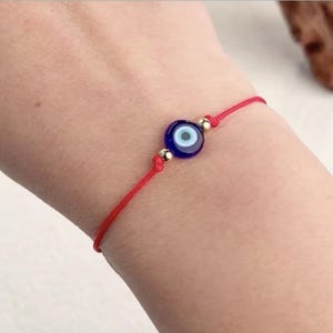 May include: Red string bracelet featuring a blue and white evil eye charm. The charm is round with a dark blue center and a light blue iris. Small gold-colored beads accent the charm. The bracelet is tied with a sliding knot.