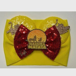 May include: Yellow headband with a red bow and a gold glitter lion king design. The design includes the words "Hakuna Matata".