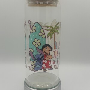 May include: A clear glass can-shaped tumbler with a bamboo lid. The tumbler features a colourful graphic of Lilo and Stitch on a beach scene with a surfboard, palm tree, and kite. The tumbler sits on a silver base.