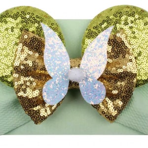 Puede incluir: Una diadema verde claro con un gran lazo dorado y blanco y dos orejas de lentejuelas. El lazo tiene un diseño de mariposa en el centro.