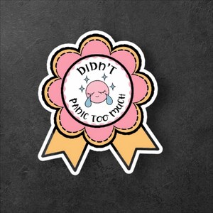 以下が含まれることがあります： 「Didn't Panic Too Much」の文字が入ったピンクと黄色のステッカー。白い円の中に泣いている顔があり、ピンクの花の形と黄色のリボンで囲まれています。