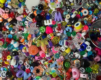 Puede incluir: Un gran surtido de cuentas, abalorios y letras de plástico de colores vivos. Las cuentas son de diversas formas, tamaños y colores, incluyendo rojo, azul, verde, amarillo, rosa y morado. También hay algunos abalorios de metal, como una carita sonriente, una estrella y un corazón. Las letras son de varios colores y tipos de letra.