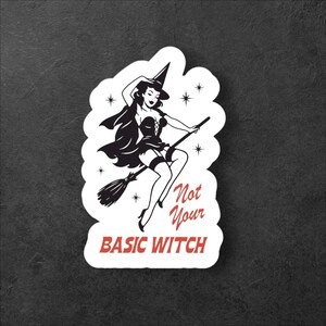 Peut inclure: Autocollant noir et blanc avec une illustration rétro d'une sorcière sur un balai. La sorcière porte un chapeau et un corset. Le texte rouge dit "Not Your Basic Witch".