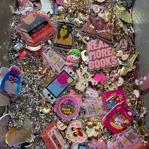 May include: A collection of colourful charms and keychains featuring various designs, including books, a video game console, a cartoon character, and a pink heart. The charms are made of different materials, including metal, plastic, and acrylic.
