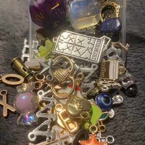 May include: A clear plastic container filled with a variety of charms and trinkets, including a purple and gold dice, a silver charm with the text "The World", a silver charm with the text "Coffee", a blue and white evil eye charm, a gold heart charm, a silver cross charm, and an orange starfish charm.