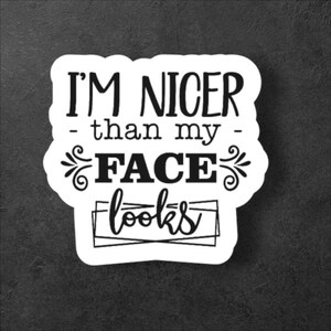 Peut inclure: Autocollant blanc avec le texte "I'M NICER - than my - FACE looks" en noir, dans une police stylisée. L'autocollant a une forme arrondie et irrégulière, sur fond gris foncé. La phrase est humoristique.