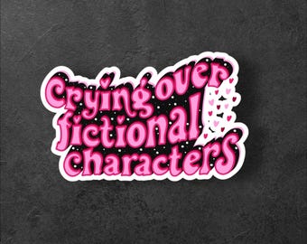 Puede incluir: Una pegatina con el texto "Crying over fictional characters" en rosa, delineado en un rosa más oscuro, sobre un fondo blanco. El fondo del texto es negro con puntos blancos y corazones rosas. La pegatina está sobre una superficie gris oscuro.