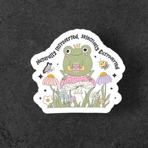 May include: A sticker featuring a cartoon frog wearing a crown and holding a cup, sitting on a mushroom. The sticker has the text "Naturally Introverted, Selectively Extroverted" and includes illustrations of flowers, a butterfly, and a bee.