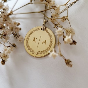 Könnte beinhalten: Goldkette mit einem runden Anhänger, der mit einem Lorbeerkranz verziert ist und die Initialen "K/A" in der Mitte eingraviert sind.