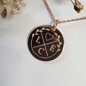 Könnte beinhalten: Eine Halskette aus Roségold mit einem runden Anhänger, auf dem ein Herz und die Buchstaben L, C und E eingraviert sind. Der Anhänger ist von einem Kranz aus Blättern umgeben.