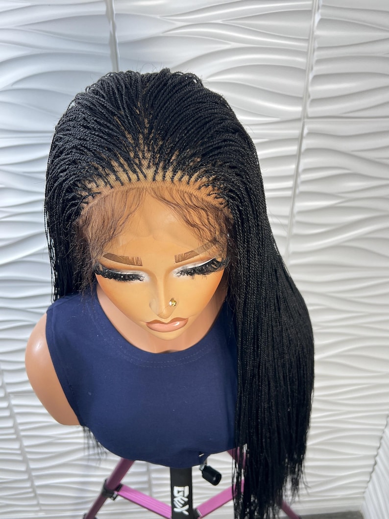 Op de afbeelding: Een zwarte gevlochten pruik met micro-vlechten, een gedefinieerde middenscheiding en een lace front basis. Het haar is lang, steil en valt over de schouders. De pruik wordt getoond op een etalagepop met een mouwloze marineblauwe top.