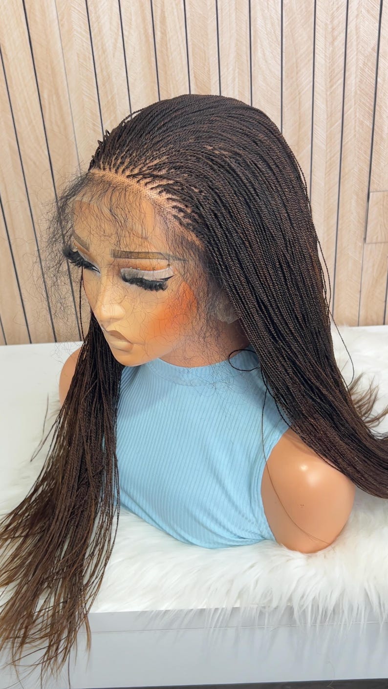 Op de afbeelding: Een donkerbruine lace front pruik met een micro-vlechttextuur. De pruik wordt getoond op een etalagepop met een lichtblauw geribbelde mouwloze top. Het haar is gestyled in lange, dunne, uniforme vlechten die over de schouders vallen.
