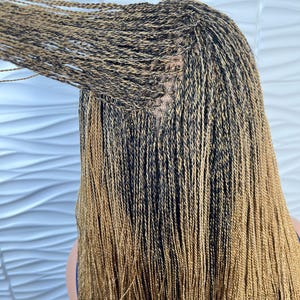 Op de afbeelding: Een pruik met lang, dun, micro-gevlochten haar in een mix van zwarte en goudbruine tinten. De vlechten zijn strak geweven vanaf de hoofdhuid, met een zichtbare kanten basis bij de kruin waar de individuele lokken aan de kap zijn bevestigd.