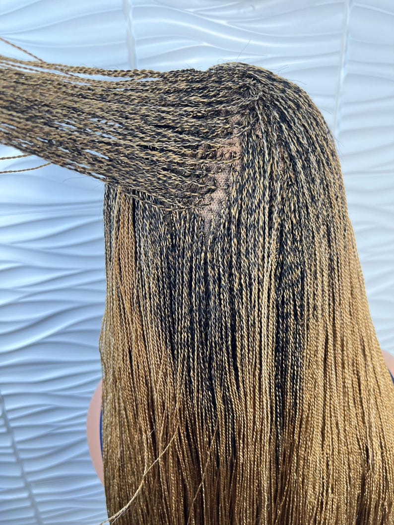 Op de afbeelding: Een pruik met lang, dun, micro-gevlochten haar in een mix van zwarte en goudbruine tinten. De vlechten zijn strak geweven vanaf de hoofdhuid, met een zichtbare kanten basis bij de kruin waar de individuele lokken aan de kap zijn bevestigd.