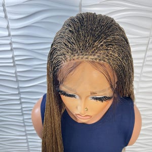 Op de afbeelding: Een lace front pruik met micro-vlechtjes in een mix van donkerbruine en goudblonde tinten. De vlechtjes zijn strak vanaf de hoofdhuid gevlochten, wat zorgt voor een natuurlijk ogende haarlijn met zichtbaar kanten gaas langs het voorhoofd.