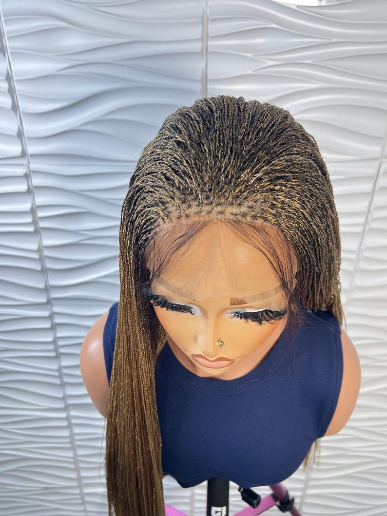Op de afbeelding: Een lace front pruik met micro-vlechtjes in een mix van donkerbruine en goudblonde tinten. De vlechtjes zijn strak vanaf de hoofdhuid gevlochten, wat zorgt voor een natuurlijk ogende haarlijn met zichtbaar kanten gaas langs het voorhoofd.