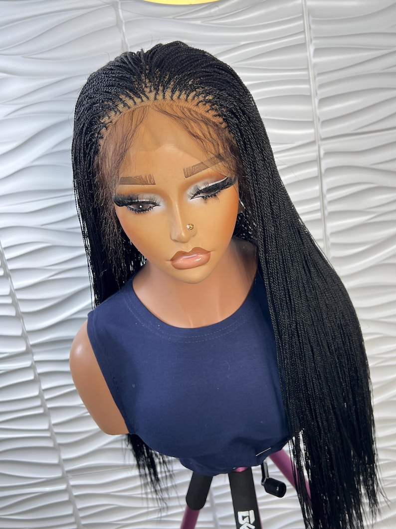 Op de afbeelding: Een lange, zwarte gevlochten pruik met micro-vlechten en een lace front haarlijn. De pruik is gestyled met een middenscheiding en valt recht tot voorbij de schouders. Het wordt getoond op een etalagepop met een donkerblauw mouwloos shirt.