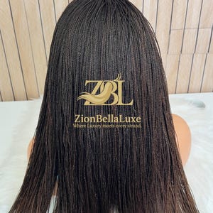 Op de afbeelding: Een donkerbruine pruik met lange, dunne, micro-gevlochten haarstrengen. De pruik wordt getoond op een etalagepophoofd en is voorzien van een gouden logo-overlay met de tekst ZBL ZionBellaLuxe Where Luxury meets every strand.