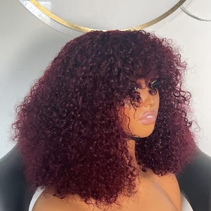 May include: A burgundy curly wig with bangs on a mannequin head. The wig is styled in a short, layered bob with loose curls.