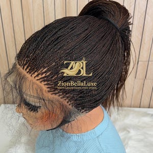 Op de afbeelding: Een donkerbruine lace front pruik gestyled in een hoge paardenstaart met micro-vlechtjes. De pruik heeft een transparante kanten haarlijn en is voorzien van de gouden tekst ZBL ZionBellaLuxe Where Luxury meets every strand op het vlechtwerk.