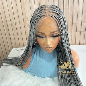 Puede incluir: Peluca trenzada gris exhibida en una cabeza de maniquí. La peluca presenta largas trenzas individuales con una línea de cabello de aspecto natural. El maniquí lleva una camiseta azul claro y está colocado sobre una superficie blanca y esponjosa. La peluca es de ZionBellaLuxe.