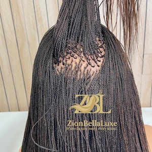 Op de afbeelding: Een donkerbruine pruik met talloze dunne, micro-gevlochten strengen gestyled in een hoge paardenstaart. De basis toont een zichtbaar hoofdhuidgedeelte waar de vlechten zijn bevestigd. Een gouden logo leest ZBL ZionBellaLuxe Where luxury meets every strand.