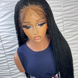 Op de afbeelding: Een lange, zwarte gevlochten pruik met micro-vlechten en een lace front haarlijn. De pruik is gestyled met een middenscheiding en valt recht tot voorbij de schouders. Het wordt getoond op een etalagepop met een donkerblauw mouwloos shirt.