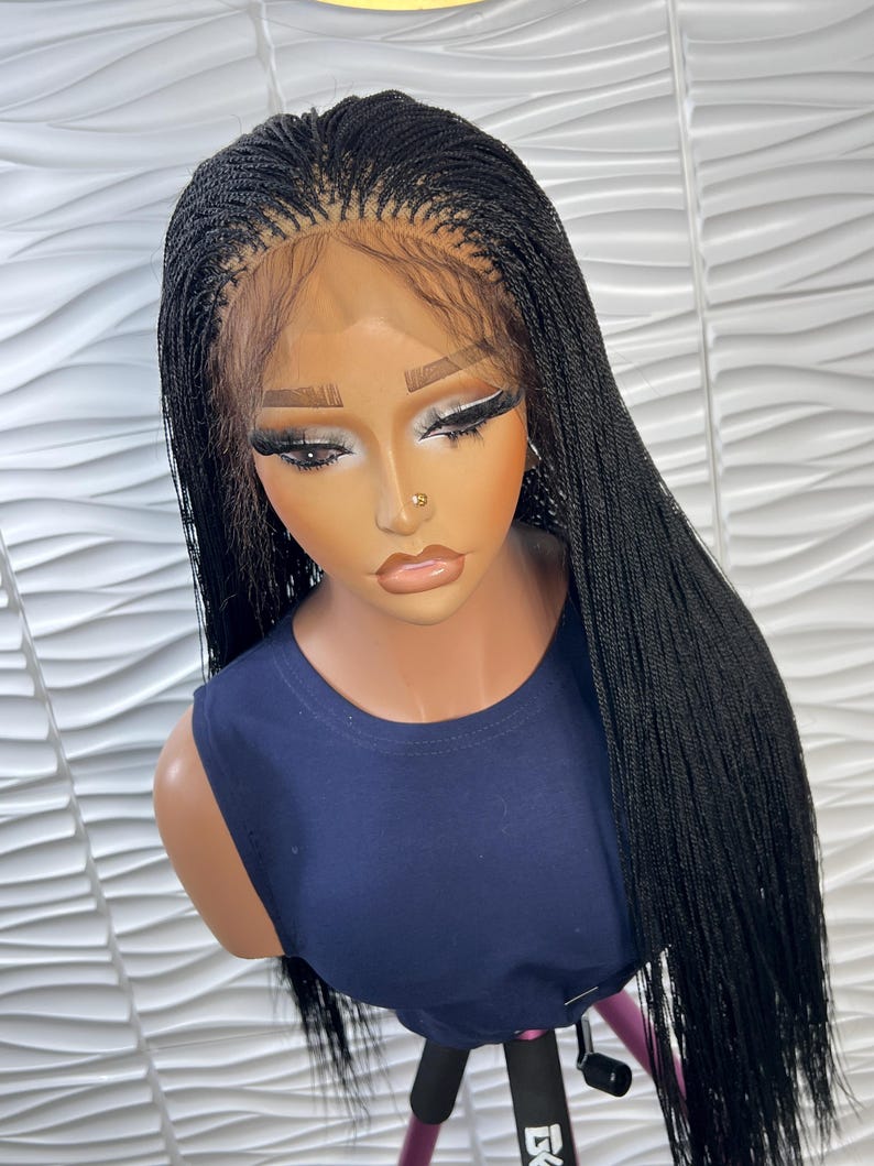 Op de afbeelding: Een lange, zwarte gevlochten pruik met micro-vlechten en een lace front haarlijn. De pruik is gestyled met een middenscheiding en valt recht tot voorbij de schouders. Het wordt getoond op een etalagepop met een donkerblauw mouwloos shirt.