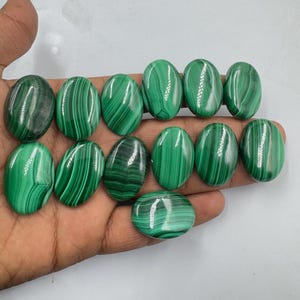 Op de afbeelding: Een verzameling ovaalvormige malachiet edelstenen, met een levendige groene kleur en kenmerkende donkergroene bandpatronen. De gepolijste stenen worden op een hand getoond, wat hun natuurlijke schoonheid en potentieel voor sieraden of decoratie benadrukt.