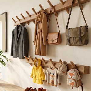 May include: Two horizontal wooden wall-mounted coat racks with multiple angled pegs. The top rack holds a grey blazer, a tan trench coat, a tan leather shoulder bag, and a green canvas messenger bag. The bottom rack holds a yellow raincoat, a beige sweater with a deer graphic, a beige knit beanie, and a fox-print backpack.