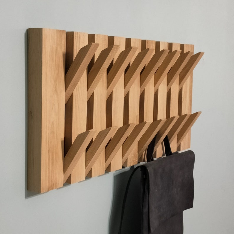 May include: A wooden wall-mounted coat rack with multiple angled hooks. The rack is made of light brown wood and has a natural finish. A black bag is hanging on one of the hooks.