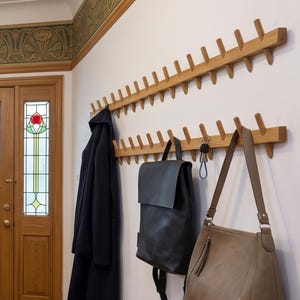 Könnte beinhalten: Zwei lange, horizontale Wandgarderoben aus Holz mit mehreren Haken und einer hellbraunen Holzmaserung. Ein dunkelblauer Mantel, ein schwarzer Lederrucksack und eine beige Umhängetasche hängen an den Haken der unteren Garderobenleiste.